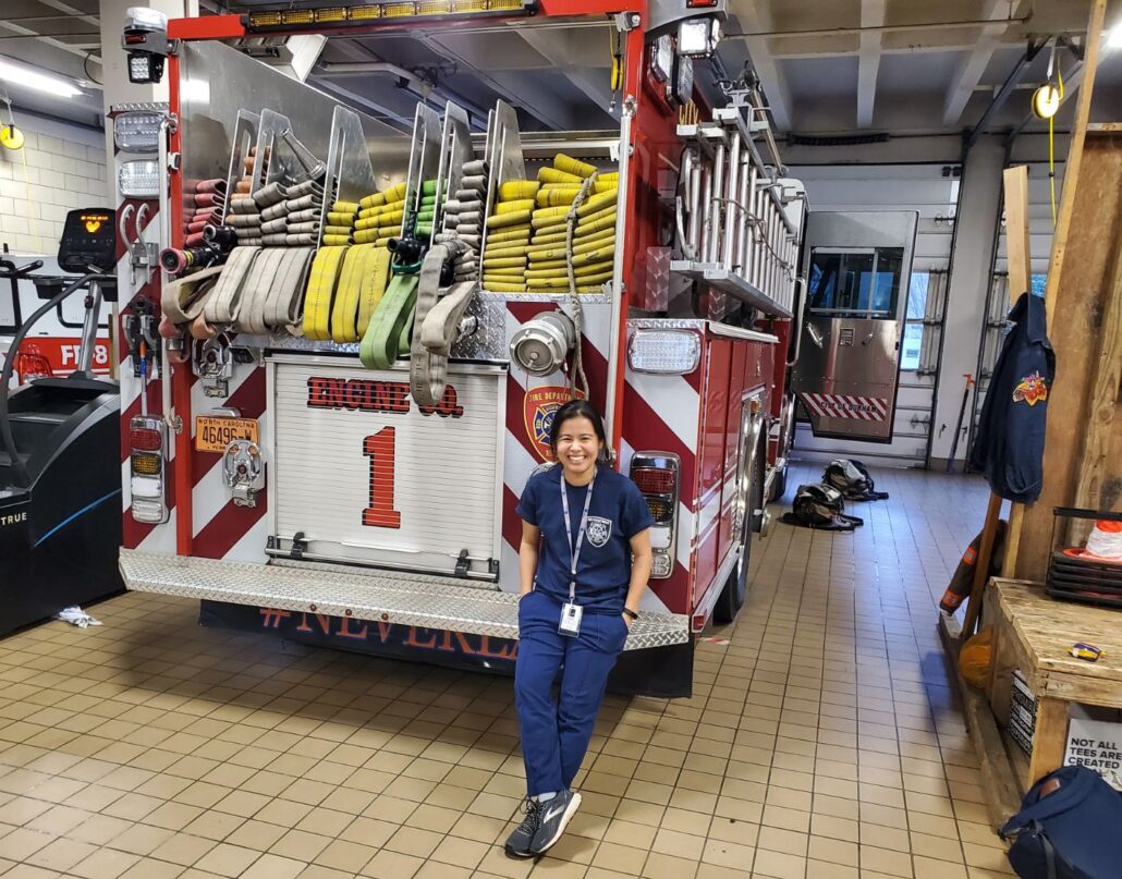 Sarah, a woman with black hair, wears a blue shirt and blue pants. She is standing in front of a fire truck and in a fire department.