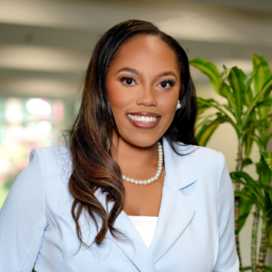Sheila, a Black woman with brown hair, wears a white shirt, pale blue suit and pearl necklace and earrings.