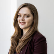 Kayla, a white woman with auburn hair, wears a paisley colored burgundy top and burgundy blazer.