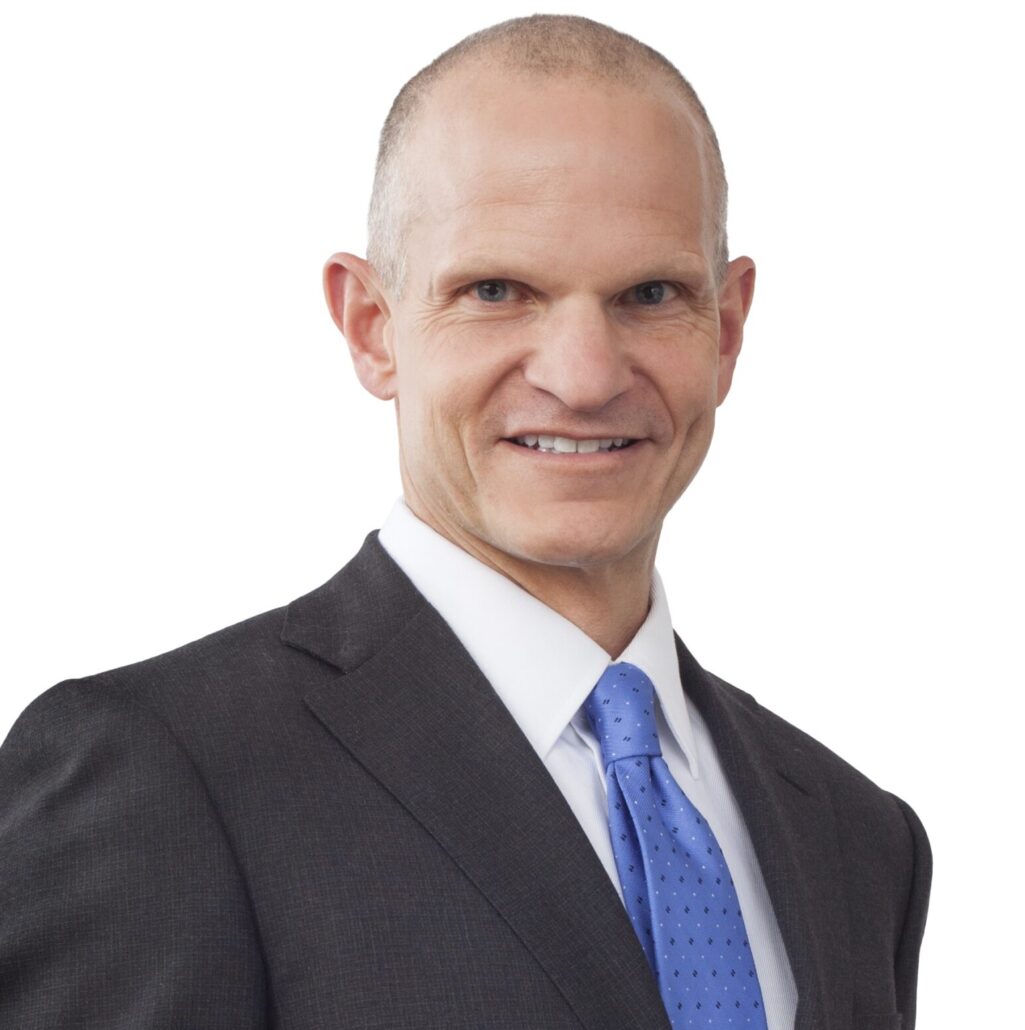 Chris, a white man with a shaved head, wears a white shirt, blue tie and black jacket.