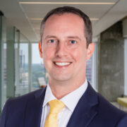 Nicholas, a white man with brown hair, wears a white shirt, yellow tie and black suit.