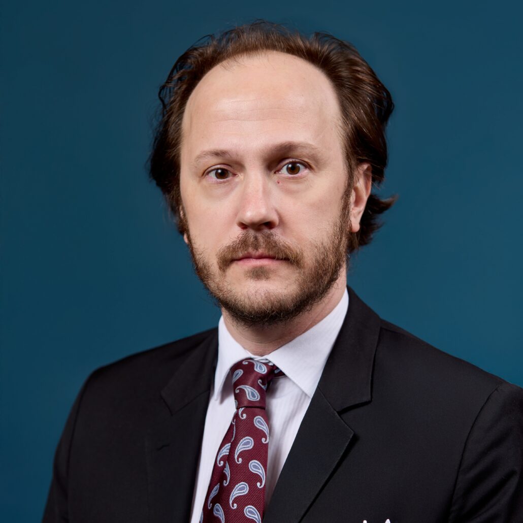 Andrew, a white man with brown hair, wears a white shirt, maroon tie with a blue paisley pattern and a black suit.