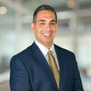 Brian, a white man with brown hair, wears a white shirt, navy suit and gold tie.