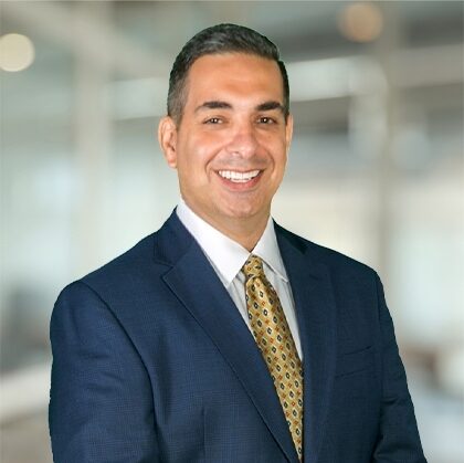 Brian, a white man with brown hair, wears a white shirt, navy suit and gold tie.