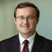 Lukas, a white man with brown hair and glasses, wears a white shirt, burgundy tie and black jacket.