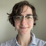Elizabeth, a white woman with short brown hair and clear glasses, wears a pale blue and white striped button-down shirt.