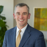Chad, a white man with grey hair, wears a white shirt, gold tie and dark grey jacket.