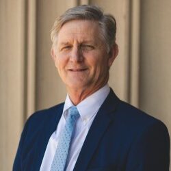 Jim, a white man with grey and brown hair, wears a white shirt, pale blue tie and navy suit.