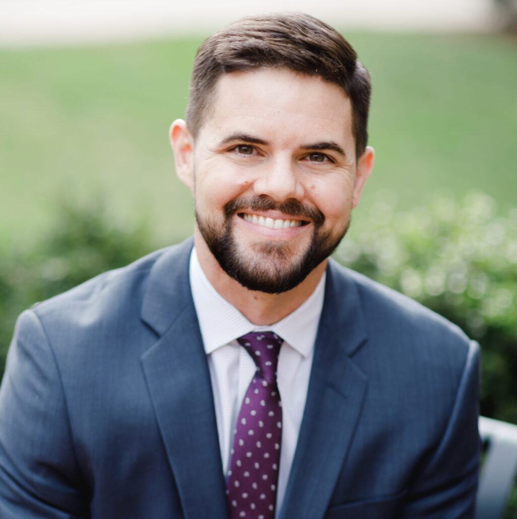 Jeremy, a white man with brown hair and a beard, wears a white shirt, navy suit and burgundy tie.