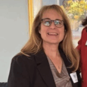 Joyce, a white woman with blond hair, wears a light grey blouse and dark grey suit.