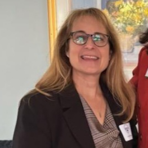 Joyce, a white woman with blond hair, wears a light grey blouse and dark grey suit.