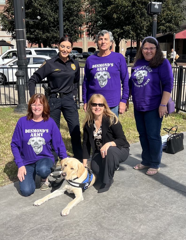 Joyce, a white woman with blond hair, is dressed in a black suit and is kneeling with her face toward the camera while her right hand is on a dog who sits on pavement. Two women and one man are also in the photo, and they are wearing purple shirts that say "Desmond's Army." A female police officer is also in the photo.