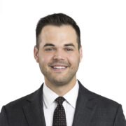 Troy, a white man with black hair, wears a white shirt, black tie and black suit.