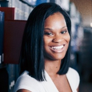 Sa'metria is a Black woman with black hair. She is wearing a white blouse and is seated in a library with an open book. 