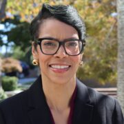 Stacey, a Black woman with black hair and black glasses, wears a black suit and is standing outside.