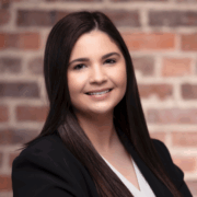 Olivia, a woman with dark brown hair, wears a white shirt and dark brown jacket. 