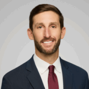 JD, a white man with brown hair and a beard, wears a white shirt, red tie and black blazer. 