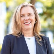 Katherine, a white woman with blond hair, wears a white blouse and dark grey suit.