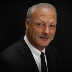 Paul, a white man with grey hair and a mustache, wears a white shirt, black tie and black blazer.