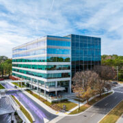 The exterior of the Glenlake Three office building is shown. It is a building with large reflective windows and there are some trees around it.