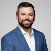 Drew, a white man with brown hair and a beard, wears a white shirt and a blue suit. 