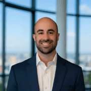 Michael, a white man with a shaved head and a dark brown beard, wears a white shirt and black blazer.