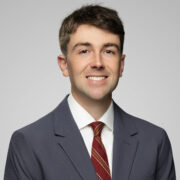 Walker, a white man with brown hair, wears a white shirt, red tie and grey suit. 