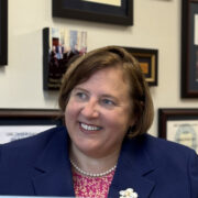 Kim, a white woman with shoulder-length brown hair, wears a pin and orange dress and blue blazer with a small off-white flower pin. She is seated in her office. 