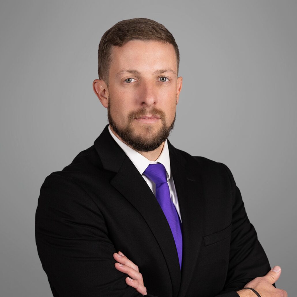 Tyler, a white man with brown hair and a beard, wears a white shirt, purple tie and black jacket.