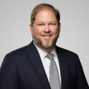 Robert, a white man with brown hair and a beard, wears a white shirt grey tie and black jacket. 