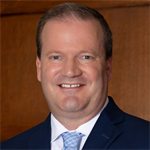 Mark, a white man with brown hair, wears a white shirt, pale blue tie and navy jacket. 