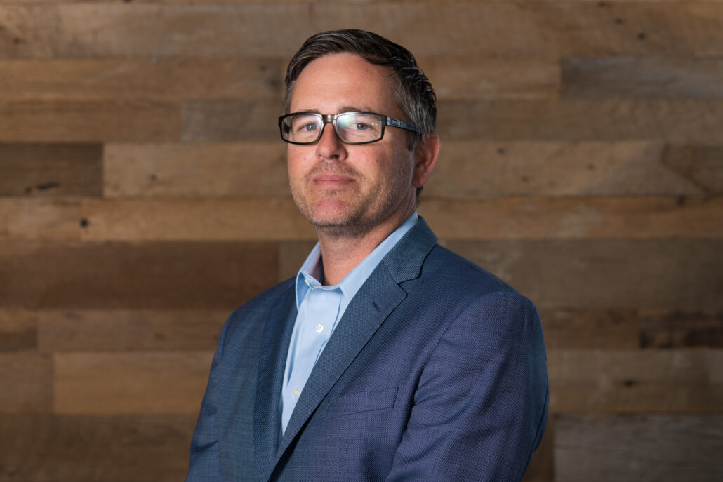 Chris, a white man with brown hair and brown glasses, wears a light blue shirt and dark grey jacket.