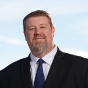 Christopher, a white man with brown hair and a beard, wears a white shirt, black blazer and blue tie.