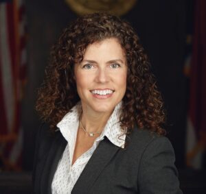 Connie, a white woman with brown hair, wears a white blouse and dark grey suit.