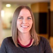 Stephanie, a white woman with light brown hair wears a dark grey dress and red necklace.