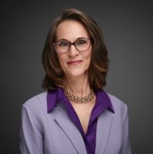Judge Debra Sasser, a white woman with brown hair and brown glasses, wears a purple blouse and pale purple jacket.