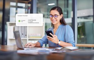 "A woman with dark hair and glasses is wearing a blue blouse. She holds a phone, and a notification appears that says, "A compliant test was received."