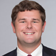 Spencer, a white man with brown hair, wears a white shirt, red tie and black jacket.
