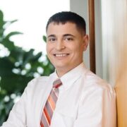 Cosmo, a white man with brown hair, wears a white shirt and grey and orange striped tie.