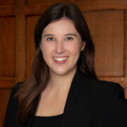 Anna, a white woman with brown hair, wears a black dress and black blazer.