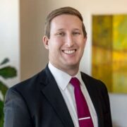 Joey, a white man with light brown hair, wears a white shirt, red tie and black suit. 