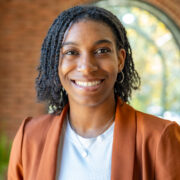 Nya, a Black woman with black hair, wears a white blouse and orange brown jacket. 