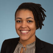 Aishaah is a Black woman with black hair. She is wearing a silver blouse with a black jacket.