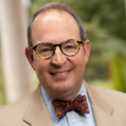 Dean Brinkley, a white man with brown hair, wears tortoiseshell-colored round glasses, a blue shirt, a tan suit, and a brown bowtie.
