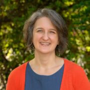 Elizabeth, a white woman with brown hair, is wearing a blue blouse and orange sweater. 