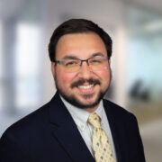 Christian, a white man with black hair and a beard and mustache, wears a pale grey shirt, yellow tie and black jacket.