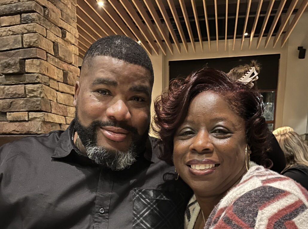 LJ, a Black woman with curly dark brown hair, wears a grey and cream sweater. Her husband is a Black man with black hair and beard who is wearing a black button-down shirt.