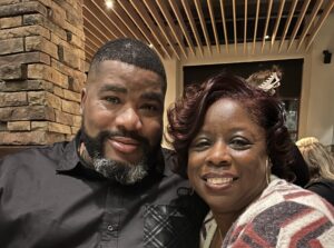 LJ, a Black woman with curly dark brown hair, wears a grey and cream sweater. Her husband is a Black man with black hair and beard who is wearing a black button-down shirt.