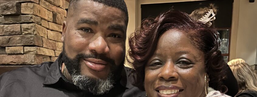 LJ, a Black woman with curly dark brown hair, wears a grey and cream sweater. Her husband is a Black man with black hair and beard who is wearing a black button-down shirt.