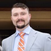 Joshua, a white man with brown hair and a beard, wears a blue shirt, grey suit and orange and white and blue striped tie.